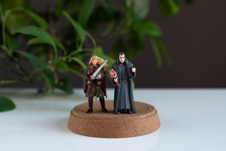 May include: Two miniature figurines on a cork base. One figurine is a woman with long blonde hair wearing a red and gold outfit and holding a sword. The other figurine is a man with black hair wearing a black robe and holding a red object.