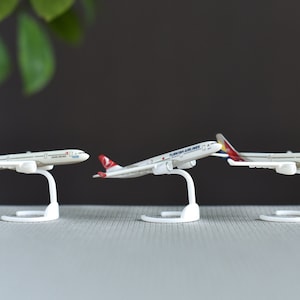 May include: Three miniature airplane models, each mounted on a white stand. The planes feature various airline liveries, including red, white, and blue designs. The models are displayed against a neutral background.