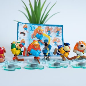 May include: A set of seven colourful plastic hockey player figurines, each with a unique design and holding a hockey stick. The figurines are arranged on a white surface with blue and green accents.