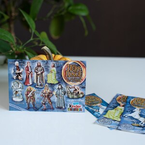 May include: A blue and gold card with images of characters from The Lord of the Rings. The card says "The Herr der Ringe" and "Die Abenteuer beginnen" in German. The card also says "Kinder &Uuml;berraschung" and has a picture of a Kinder egg. There are three smaller blue and gold cards with images of characters from The Lord of the Rings.