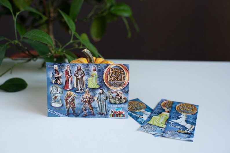 May include: A blue and gold card with images of characters from The Lord of the Rings. The card says "The Herr der Ringe" and "Die Abenteuer beginnen" in German. The card also says "Kinder &Uuml;berraschung" and has a picture of a Kinder egg. There are three smaller blue and gold cards with images of characters from The Lord of the Rings.