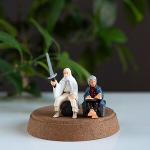 May include: A white-bearded figure in a white robe holds a sword, while another figure in a blue robe sits and reads a book. Both figures are on a round, brown base.