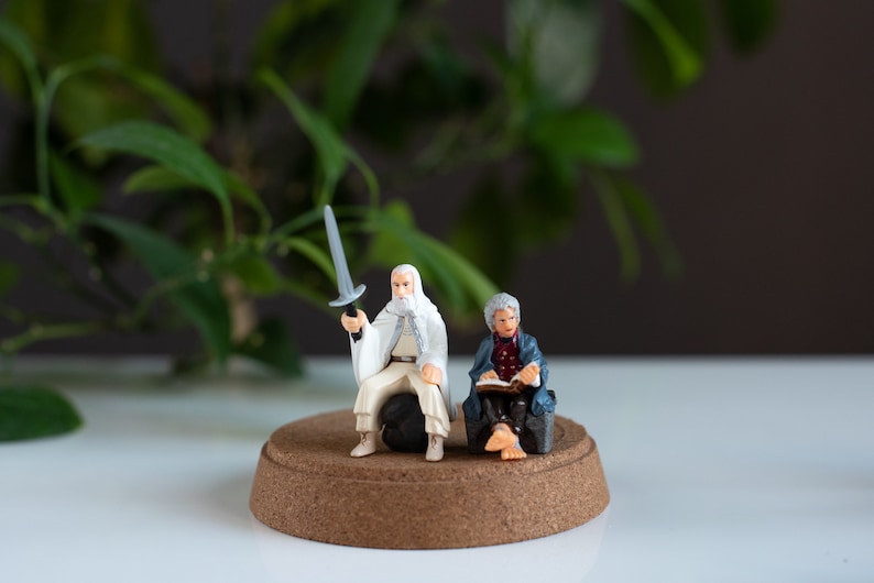 May include: A white-bearded figure in a white robe holds a sword, while another figure in a blue robe sits and reads a book. Both figures are on a round, brown base.