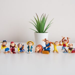 Pode incluir: Uma coleção de 11 figurinhas de macaco de plástico coloridas, algumas segurando bananas, outras com chapéus e roupas. As figurinhas estão dispostas em fila sobre uma superfície branca.