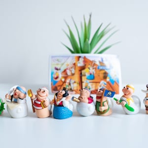 Può includere: Un set di nove figurine di plastica colorate di pirati dei cartoni animati, ognuna con un'espressione diversa e che tiene un oggetto diverso. Le figurine sono disposte in fila su una superficie bianca.