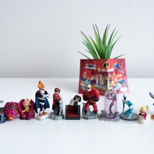 Puede incluir: Un conjunto de 10 figuras de plástico de la película de Disney Pixar "Los Increíbles". El conjunto incluye a Mr. Increíble, Elastigirl, Violet, Dash, Síndrome, Frozone y la mascota de la familia Increíble, un perro llamado 'Bolt'. Las figuras están todas en sus trajes de superhéroes y están en poses de acción. El conjunto también incluye un pequeño coche de plástico y un pequeño avión de plástico.