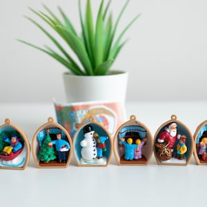 May include: Six miniature Christmas ornaments with various winter scenes, including a snowman, Santa Claus, and children. Each ornament is encased in a wooden-colored frame with a blue interior. A small potted plant is in the background.
