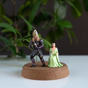 May include: A miniature figurine set featuring a knight in black armor with a sword and a woman in a green dress. The figurines are standing on a cork base.