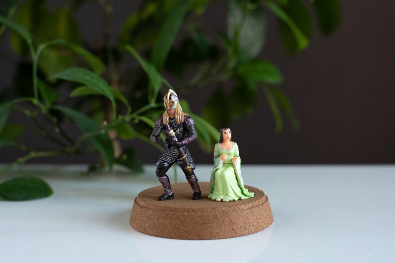 May include: A miniature figurine set featuring a knight in black armor with a sword and a woman in a green dress. The figurines are standing on a cork base.