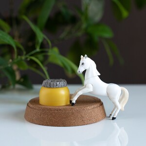 May include: A white plastic toy horse standing on a cork base with a yellow and gray plastic container in front of it.
