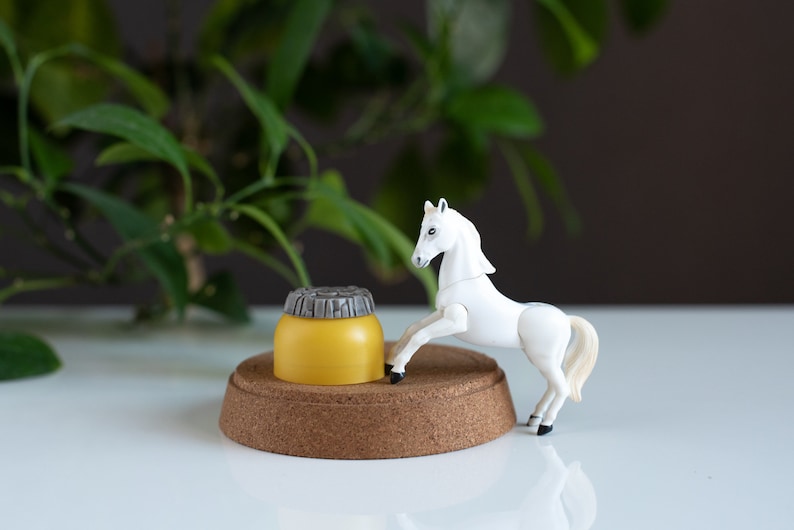 May include: A white plastic toy horse standing on a cork base with a yellow and gray plastic container in front of it.