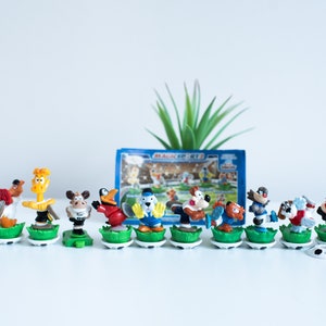Può includere: Un set di 10 figurine di plastica di animali dei cartoni animati che indossano divise da calcio. Le figurine sono in piedi su basi verdi con palloni da calcio bianchi. Le figurine fanno parte di una collezione Magic Sets.