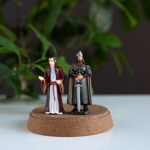 May include: Two miniature figurines on a cork base. The figurine on the left is wearing a long, white and red robe with a blue sash. The figurine on the right is wearing a silver and black armor with a sword.