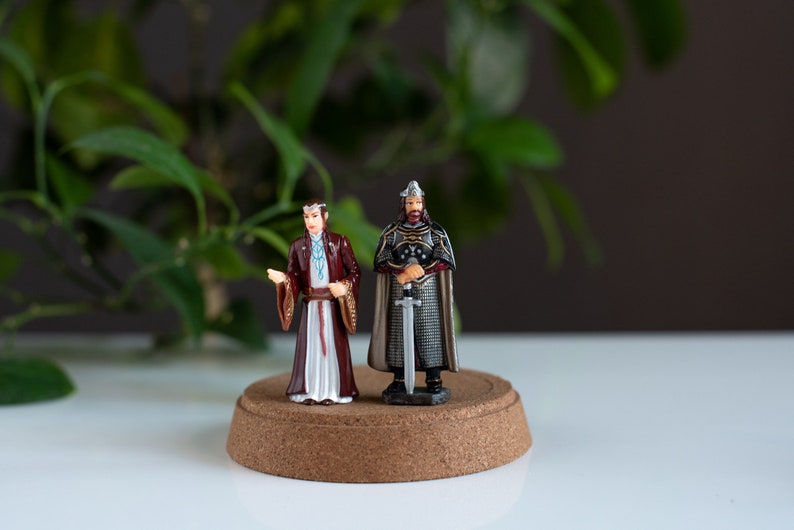 May include: Two miniature figurines on a cork base. The figurine on the left is wearing a long, white and red robe with a blue sash. The figurine on the right is wearing a silver and black armor with a sword.