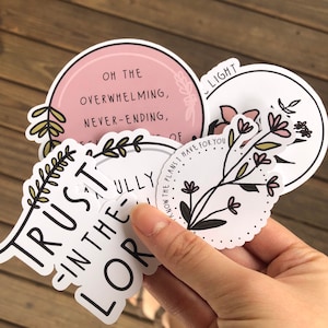 May include: A set of five round stickers with inspirational quotes and floral designs. The stickers feature phrases like "Trust in the Lord" and "Oh the overwhelming, never-ending..."  The stickers are white with black text and floral illustrations.