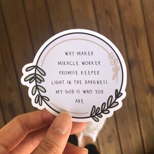 May include: A white round sticker with a brown circle in the center. The text inside the circle reads "WAY MAKER, MIRACLE WORKER, PROMISE KEEPER, LIGHT IN THE DARKNESS, MY GOD IS WHO YOU ARE". The sticker is decorated with green leafy branches.