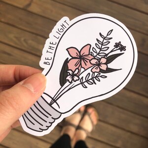 May include: A white lightbulb sticker with a black outline. Inside the lightbulb is a bouquet of pink flowers with green leaves. The text "BE THE LIGHT" is written on the side of the lightbulb.