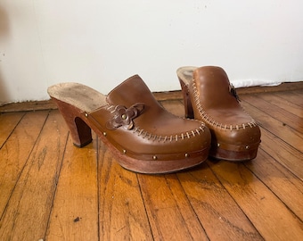 May include: A pair of brown leather platform clogs with a flower detail on the top. The clogs have a high heel and a stitched design around the edge.