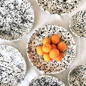 Könnte beinhalten: Ein Set weißer Keramikschalen mit schwarzem Spritzmuster. Die Schalen sind in verschiedenen Größen und auf einer weißen Oberfläche angeordnet. Eine Schale in der Mitte des Bildes ist mit frischen Aprikosen gefüllt.