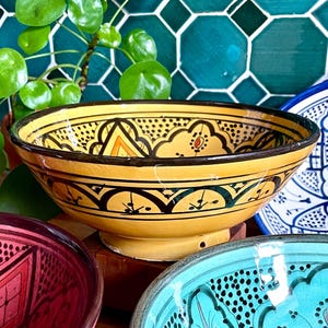 Könnte beinhalten: Drei Keramikschalen mit aufwendigen Designs. Die Schalen sind gelb, rosa und türkis mit schwarzen und weißen Details. Die Schalen stehen auf einer Holzoberfläche.