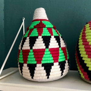 K&ouml;nnte beinhalten: Ein geflochtener Aufbewahrungskorb mit einem geometrischen Muster in Schwarz, Wei&szlig;, Rot und Gr&uuml;n. Der Korb ist rund und hat eine spitze Oberseite.