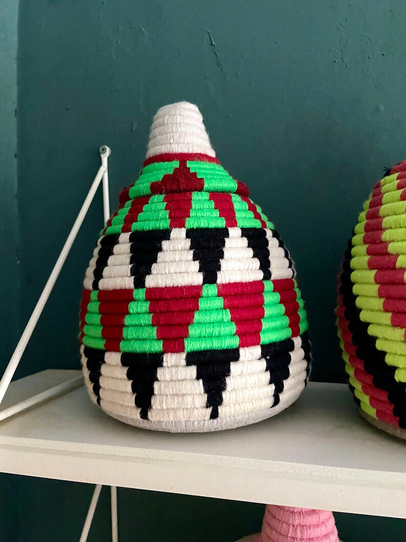 K&ouml;nnte beinhalten: Ein geflochtener Aufbewahrungskorb mit einem geometrischen Muster in Schwarz, Wei&szlig;, Rot und Gr&uuml;n. Der Korb ist rund und hat eine spitze Oberseite.