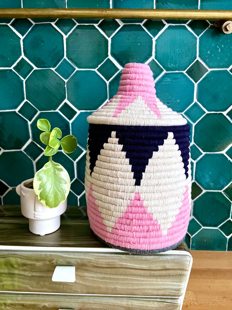 K&ouml;nnte beinhalten: Ein geflochtener Aufbewahrungskorb mit einem geometrischen Muster in Pink, Wei&szlig; und Schwarz. Der Korb steht auf einer Holzoberfl&auml;che.