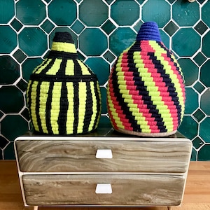 K&ouml;nnte beinhalten: Zwei geflochtene K&ouml;rbe mit schwarzen, gelben, roten und blauen Streifen stehen auf einer Kommode aus Holz mit zwei Schubladen. Die Kommode hat wei&szlig;e Schubladenkn&ouml;pfe.