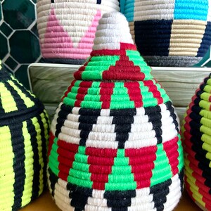 K&ouml;nnte beinhalten: Ein wei&szlig;er, gr&uuml;ner, roter und schwarzer geflochtener Korb mit konischer Form. Der Korb besteht aus einem dicken, strukturierten Material und hat ein geometrisches Muster.
