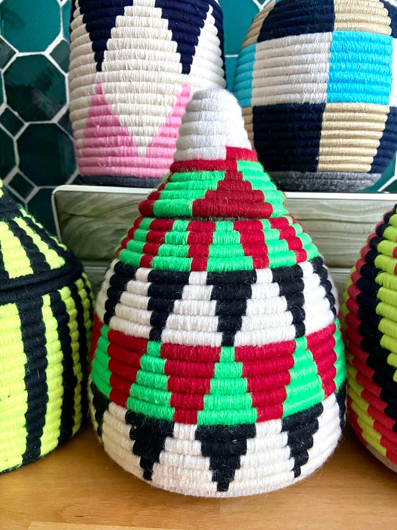 K&ouml;nnte beinhalten: Ein wei&szlig;er, gr&uuml;ner, roter und schwarzer geflochtener Korb mit konischer Form. Der Korb besteht aus einem dicken, strukturierten Material und hat ein geometrisches Muster.