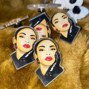 Könnte beinhalten: Eine Sammlung von Acryl-Anstecknadeln mit einem stilisierten Porträt einer Frau mit rotem Lippenstift, schwarzen Handschuhen und einem schwarzen Oberteil. Die Anstecknadeln liegen auf einer goldfarbenen, strukturierten Oberfläche. Andere Anstecknadeln mit abstrakten Formen sind ebenfalls sichtbar.
