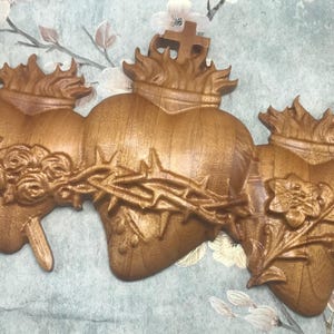 May include: Three wooden heart sculptures, each with a different design. The first heart is pierced by a sword and surrounded by roses. The second heart is adorned with a crown of thorns. The third heart is surrounded by a leafy branch.