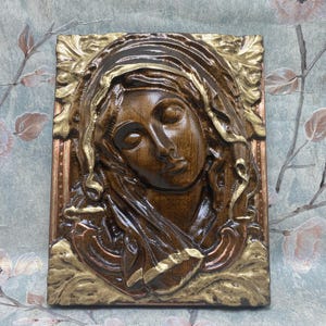 May include: A rectangular, wooden relief sculpture depicting a serene face with closed eyes, framed by a detailed headdress and ornate gold accents. The sculpture is primarily brown with gold and copper highlights, showcasing a classical theme.