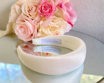 May include: A white padded headband with the text "KURY Z Collection" on a label, resting on a reflective surface. A bouquet of pink and cream-colored roses is in the background, creating a soft, romantic aesthetic.