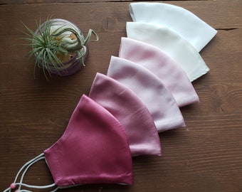 May include: Set of six satin face masks in a gradient of pink and white colors. The masks are arranged in a stack, with the darkest pink mask at the bottom and the white mask at the top.