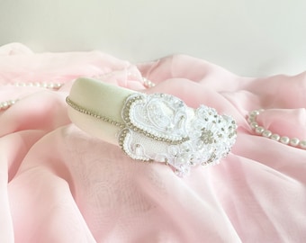Op de afbeelding: Een wit hoofdband met een groot wit bloemenmotief en strass steentjes. Het hoofdband ligt op een roze stoffen achtergrond.