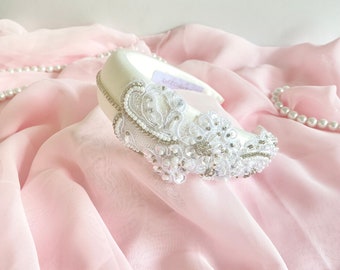 May include: A white headband with a delicate lace design and pearl embellishments. The headband is on a pink fabric background.