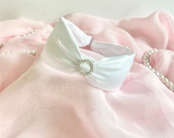 Op de afbeelding: Een wit satijnen hoofdband met een ringaccent bezet met kristallen. Het hoofdband ligt op een roze stoffen achtergrond met parelkettingen.