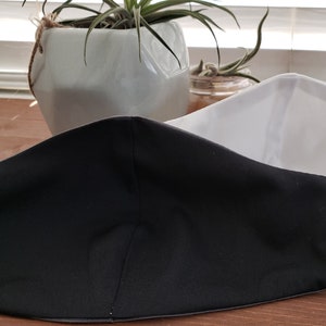 Op de afbeelding: Een zwart-wit gezichtsmasker met een witte buitenlaag en een zwarte binnenlaag. Het masker is dubbelgevouwen en ligt op een houten oppervlak.