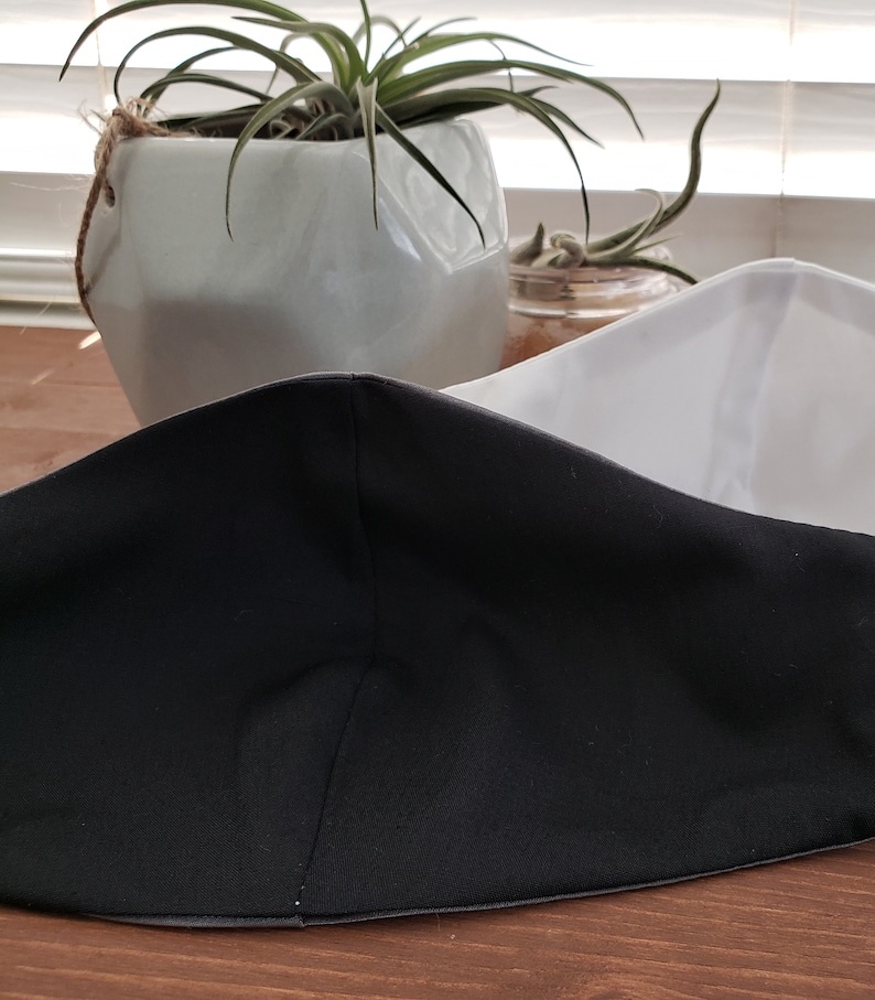 Op de afbeelding: Een zwart-wit gezichtsmasker met een witte buitenlaag en een zwarte binnenlaag. Het masker is dubbelgevouwen en ligt op een houten oppervlak.