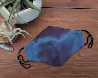 May include: A blue and purple face mask made of silk with black elastic ear loops and blue bead accents.
