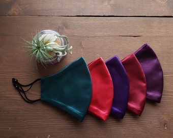 May include: Five satin face masks in different colors: teal, red, purple, burgundy, and dark purple. The masks are arranged in a row, with the teal mask at the front and the dark purple mask at the back.