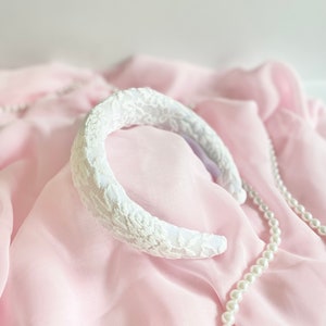May include: White lace headband on a pink fabric background with a pearl necklace.