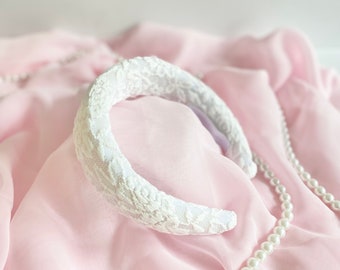 May include: White lace headband on a pink fabric background with a pearl necklace.