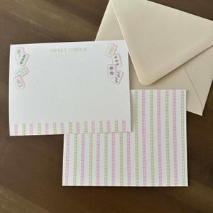 May include: Stationery set featuring a cream-colored notepad with the name "ASHLEY O'BRIEN" and Mahjong tile illustrations in pink and green. Includes a patterned card and a matching envelope.