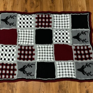 Puede incluir: Una manta de crochet con un diseño de patchwork que presenta cuadrados negros, blancos, rojos y grises. Algunos cuadrados tienen un patrón a cuadros, mientras que otros tienen una silueta de ciervo.