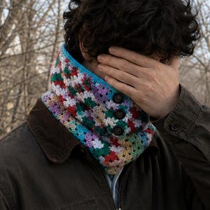 May include: A colorful crochet cowl with a button closure. The cowl features a mix of pastel colors including blue, green, pink, and white. The person is wearing a brown jacket with a corduroy collar.