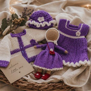 Puede incluir: Un conjunto de artículos de bebé de ganchillo hechos a mano en una cesta de mimbre. El conjunto incluye un vestido, un sombrero y un cárdigan morados con ribetes blancos, y una muñeca. Una tarjeta dice "welcome little one."