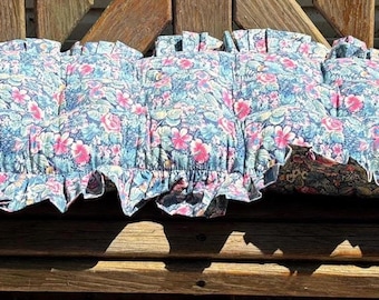 May include: A rectangular bench cushion featuring a floral pattern with pink and white flowers on a light blue background. The cushion is tufted with a ruffled edge trim and has a dark, patterned fabric visible on the underside along the front edge.