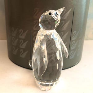 May include: A clear crystal penguin figurine with a faceted design. The penguin has a pointed beak and small black eyes. The figurine is standing in front of a gray box with swan designs.